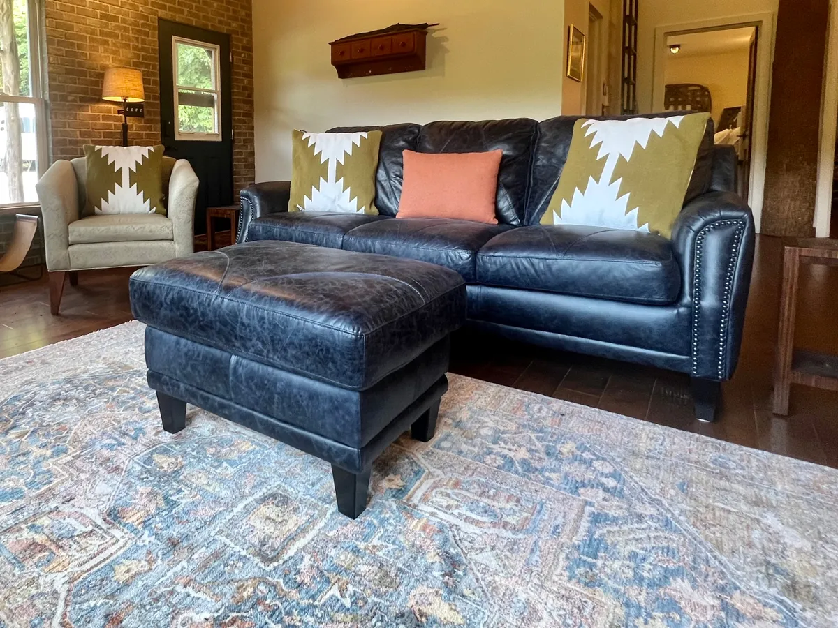 A stylish living area with a black leather sofa, colorful throw pillows, an ottoman, and a wooden side table. The space is filled with soft, natural light, perfect for relaxing or informal meetings during a workday break.