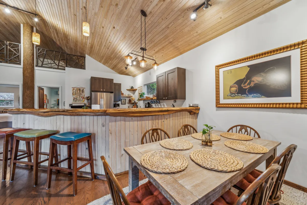 A communal dining area with a wooden table set for family dinners, woven placemats, a bar area with colorful stools, and warm ceiling details. The walls are adorned with artwork, enhancing the room's convivial, homey vibe.