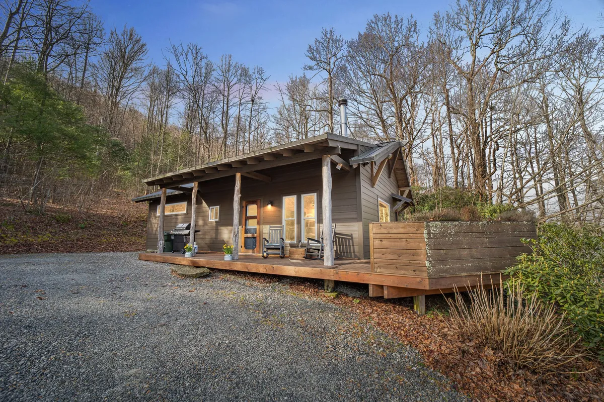The image shows a cozy wooden cabin nestled in the mountains, featuring a spacious wooden deck adorned with classic rocking chairs, a charming planter box, and large windows that invite natural light and scenic views. The deck offers a tranquil spot perfect for unwinding, with the wooded surroundings creating a peaceful, private atmosphere ideal for relaxation.