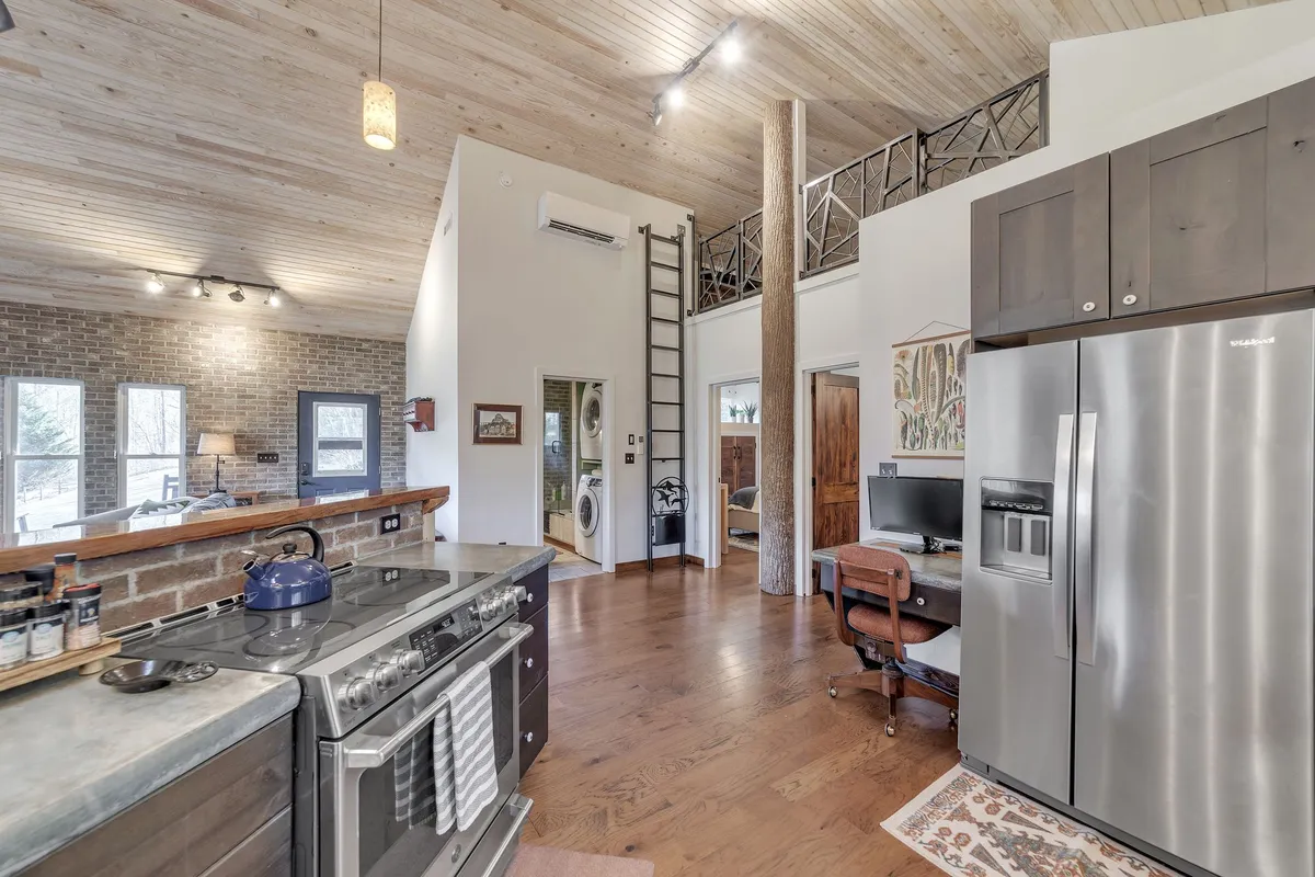 A bright, modern kitchen with a spacious island, stainless steel appliances, exposed wooden beams, and ladders leading to loft spaces. The open layout suggests opportunities for communal cooking, entertaining, and flexible group activities.