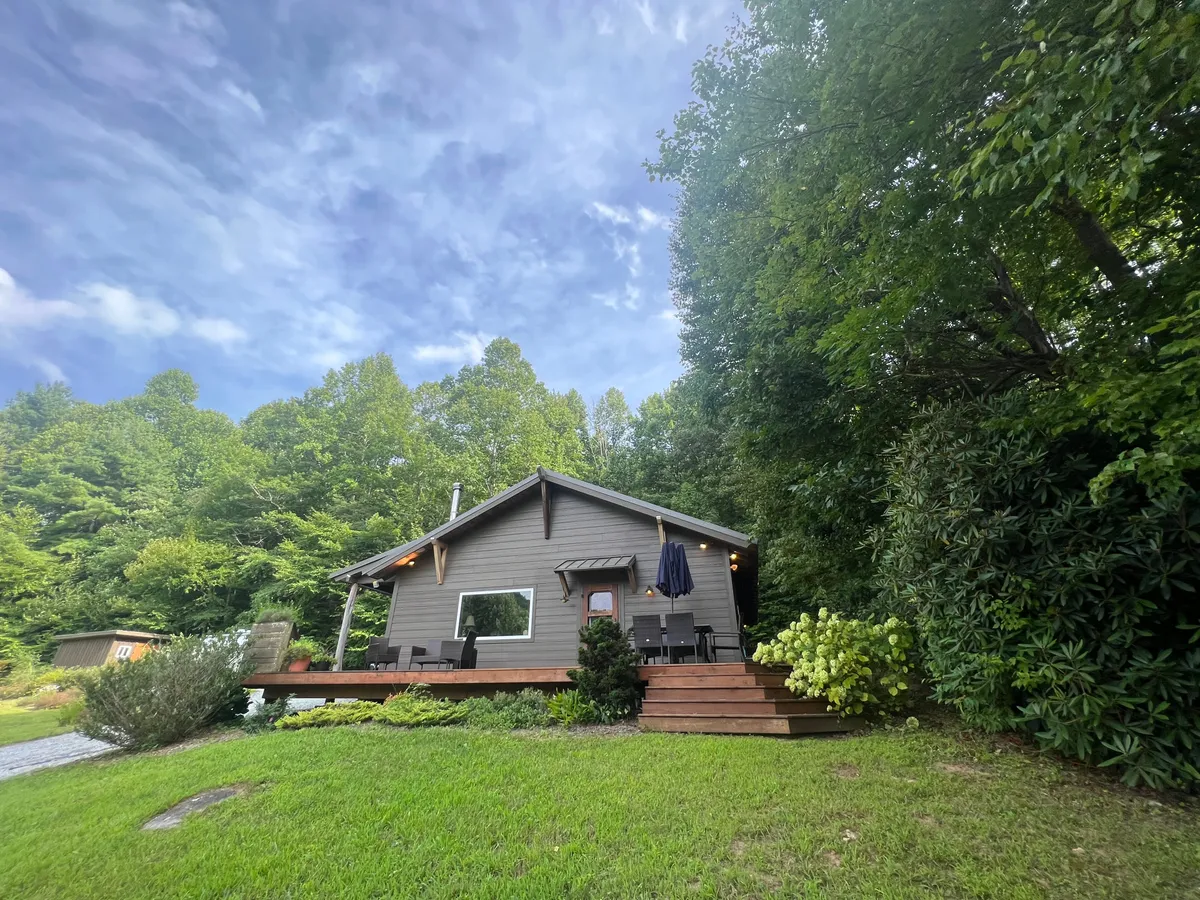 A wide exterior shot of the cabin’s deck area nestled among greenery, with the house prominently in view. The surrounding landscape suggests privacy and immersion in nature, with space for stargazing, relaxing, or setting up a picnic.