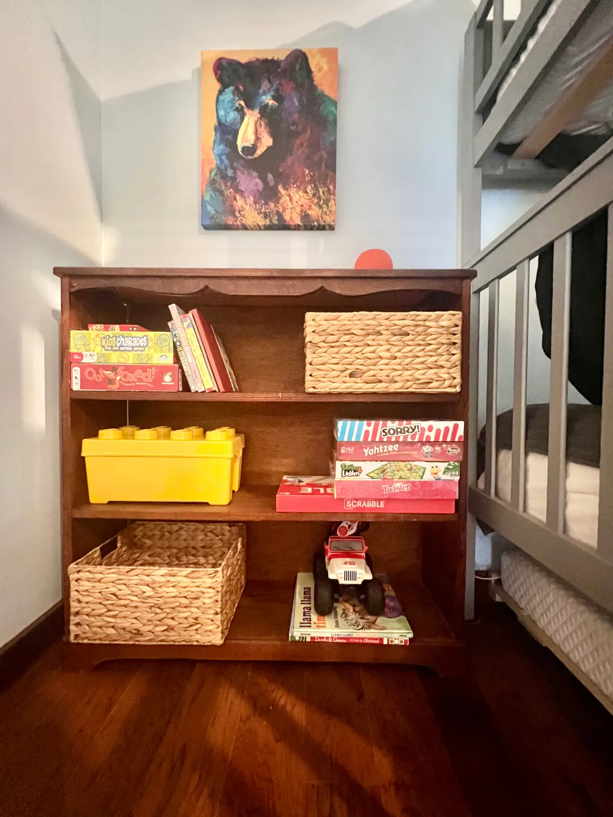 This cheerful room includes a colorful bear painting, a wooden bookshelf, a yellow storage container, and a variety of board games and toys. It radiates a playful yet relaxing vibe, inviting imaginative play and family togetherness.
