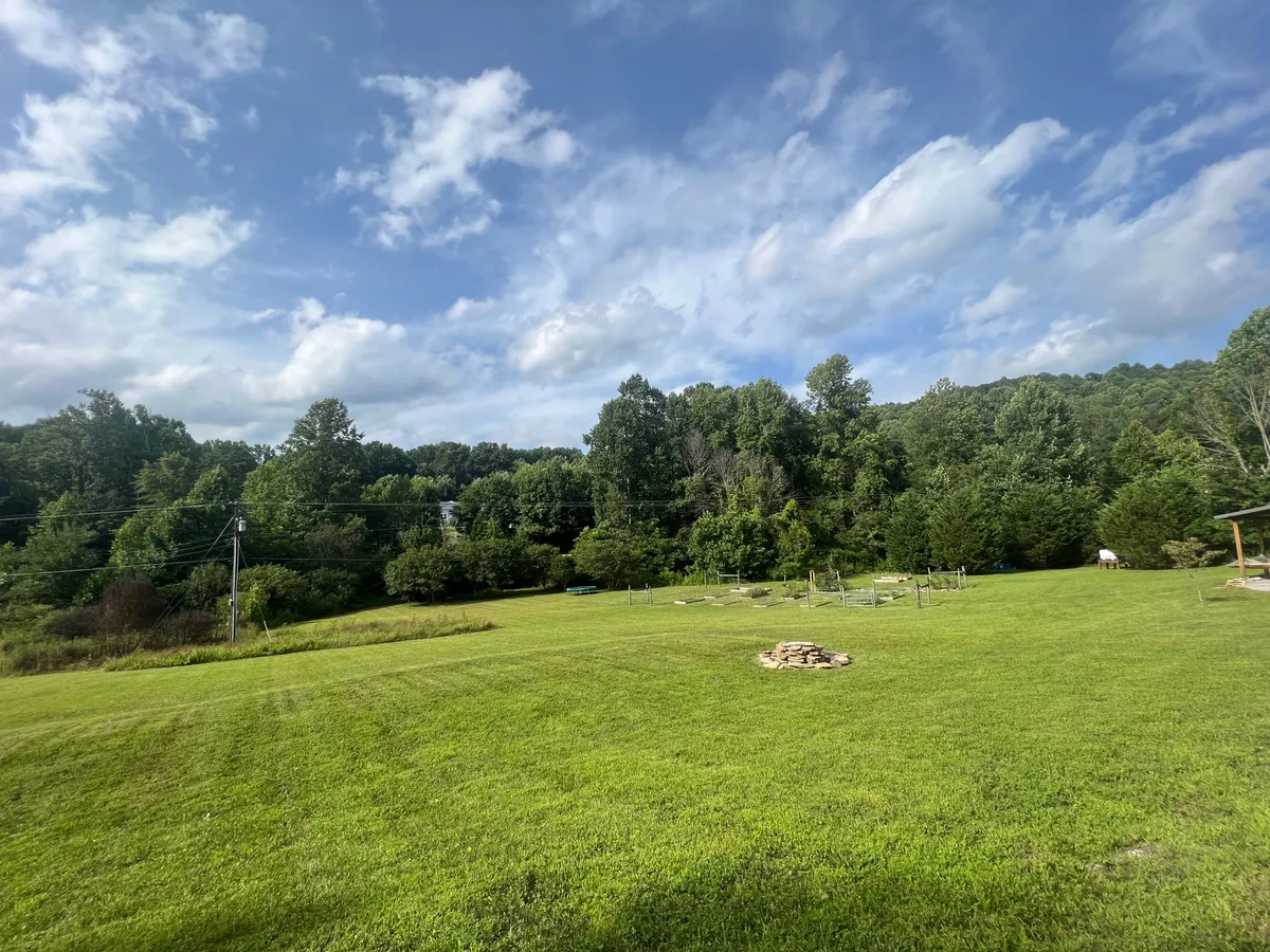 A wide, lush grassy yard stretches out toward a distant tree line, featuring a fire pit and ample open space for games or picnics. The image evokes a sense of freedom and play, ideal for families and pets, and hints at the creek and natural landscape mentioned in the text.