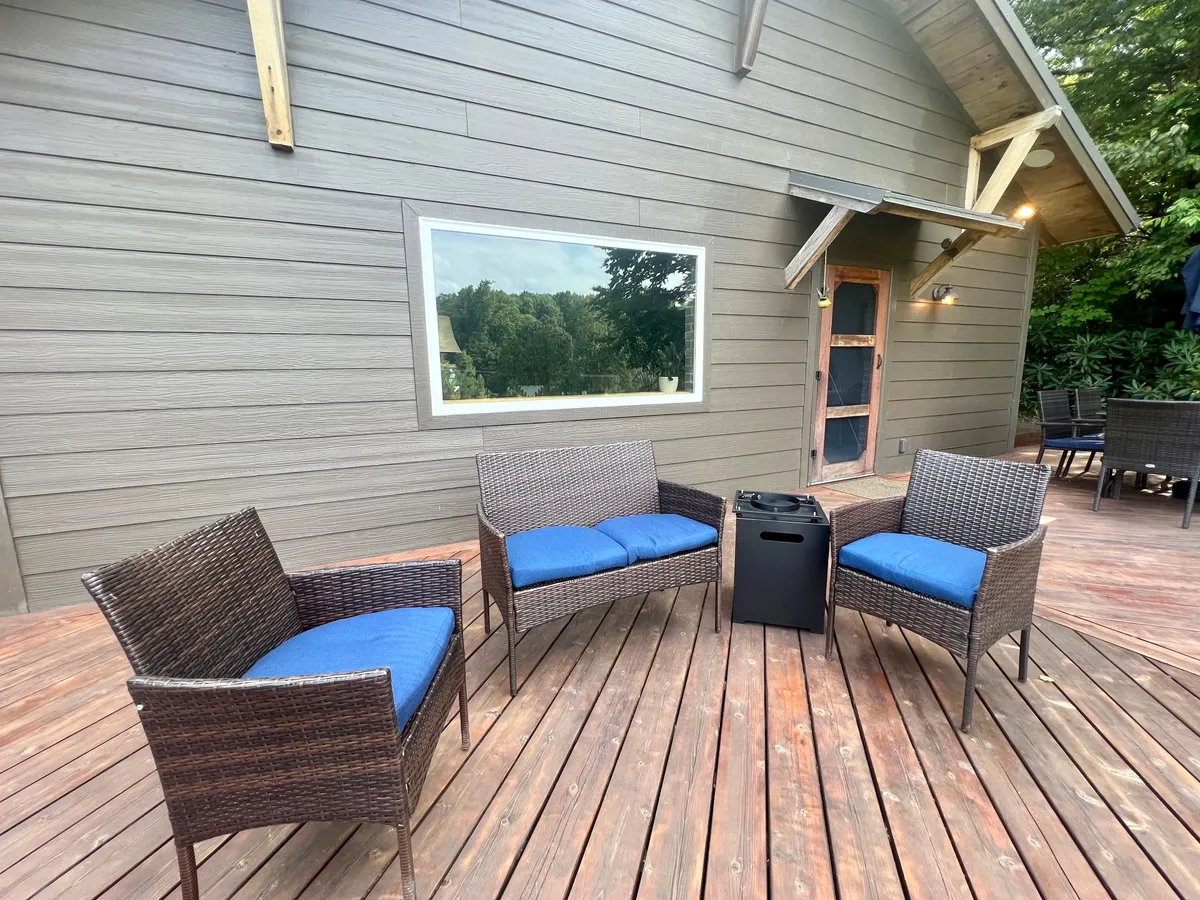 A cozy outdoor scene featuring two wicker chairs with plush blue cushions, a small table between them, and a spacious wooden deck. Sunlight pours in, offering a serene vantage point for enjoying the surrounding landscape or intimate conversation.