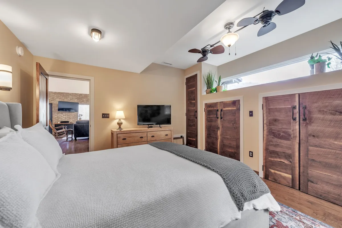This image showcases a serene bedroom with a comfortable bed, a wooden dresser, a television, closet doors, and some indoor plants. Soft natural light streams into the space, creating a tranquil atmosphere ideal for relaxing or unwinding after a long day.
