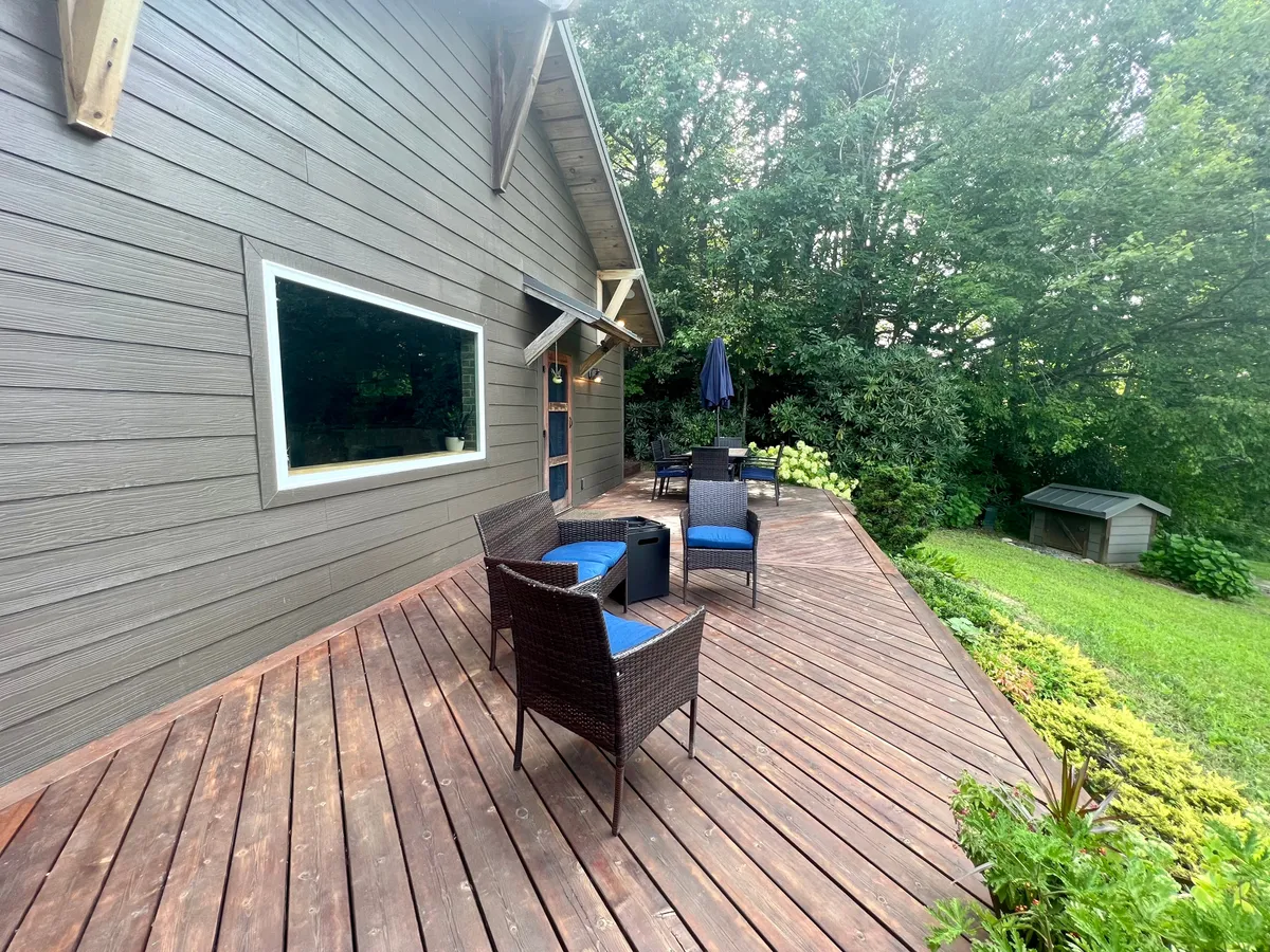 An inviting exterior deck with a pair of chairs and a table overlooking lush greenery. Natural light floods the area, and the setup suggests a tranquil space for enjoying morning coffee or evening wine. The window reflects the surrounding nature, blurring the boundaries between indoor comfort and outdoor serenity.