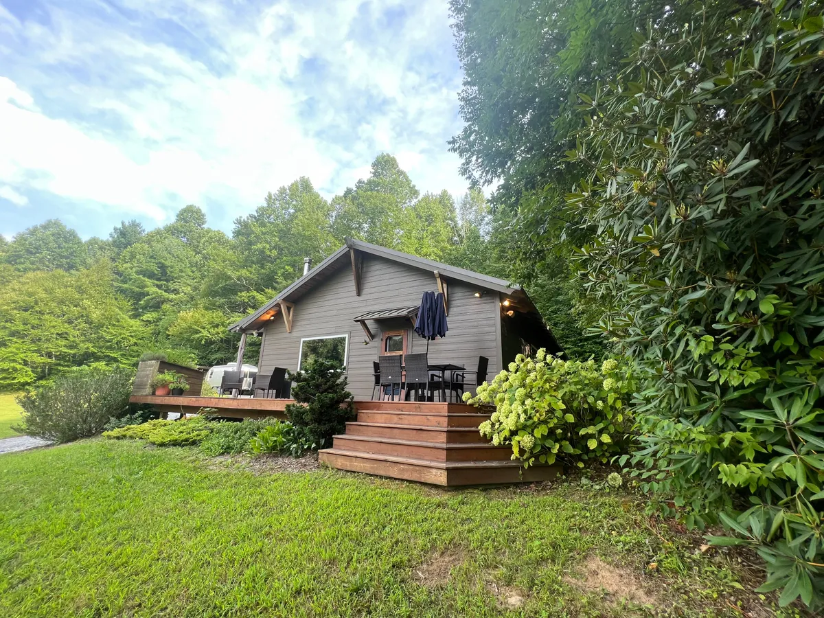 An inviting wooden deck framed by lush green foliage, featuring comfortable outdoor seating and a view of the house exterior. The setting suggests a space ideal for group gatherings, meals, or simply relaxing while surrounded by nature.