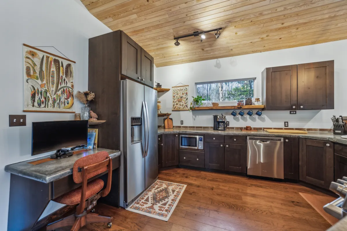 This image captures a spacious open-concept area with a warm wooden ceiling, a modern kitchen island, stylish appliances, decorative artwork, and a visible workstation nestled nearby. The scene conveys a cozy, functional space ideal for cooking, entertaining, and working from home, with abundant natural light streaming through large windows.