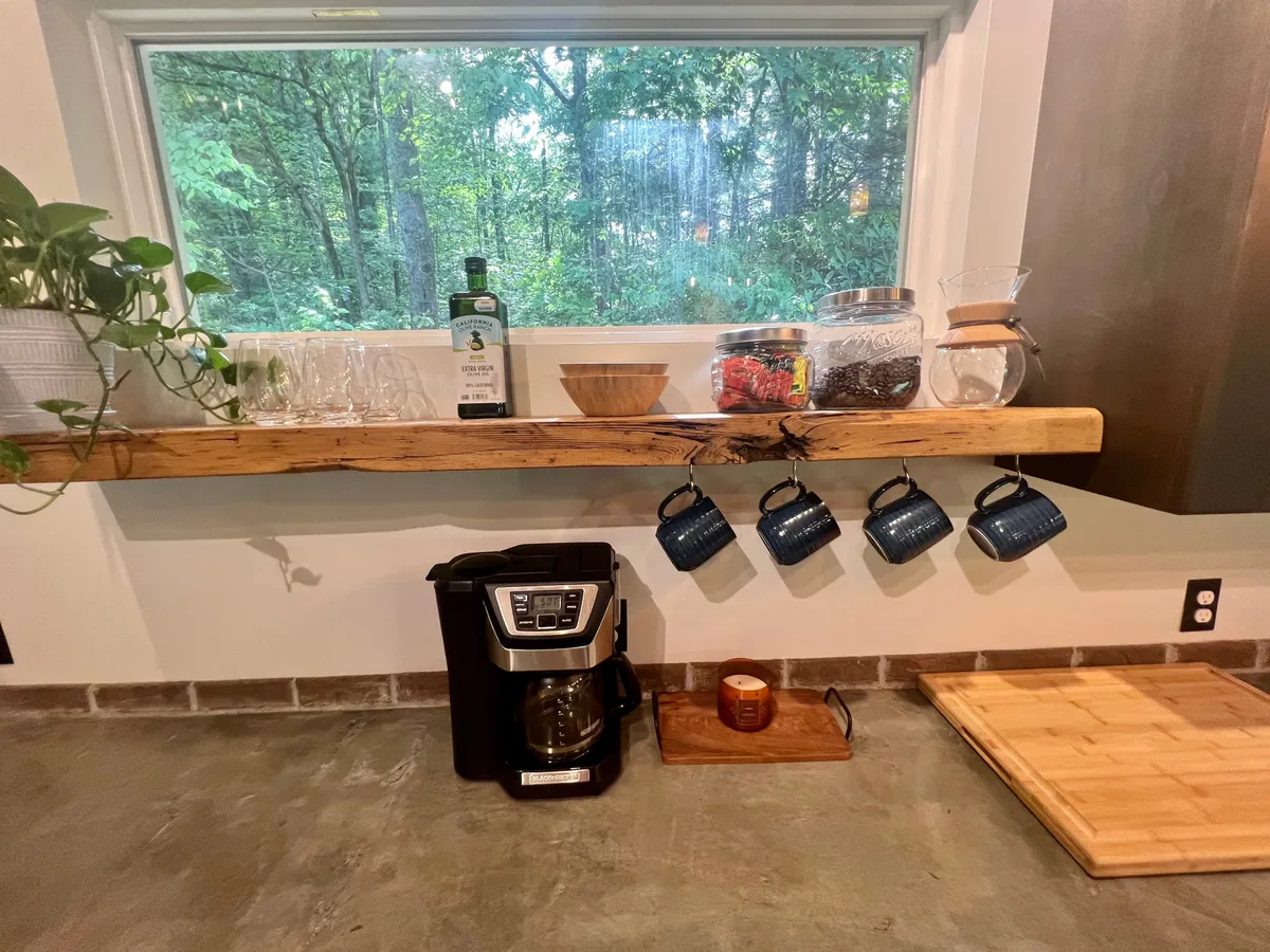 A cozy kitchen corner with a window framing mountain views, a wooden shelf, a coffee maker, and mugs hanging nearby. Morning light pours in, creating an inviting atmosphere for sipping coffee or starting the day productively.